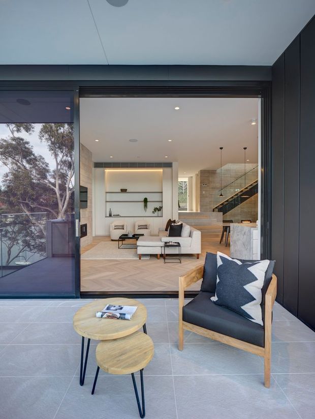 Contemporary open-plan living room viewed from a covered outdoor terrace through large sliding glass doors. The interior features pale herringbone timber flooring, a built-in illuminated shelving niche, neutral sofas and armchairs, a tiled fireplace or feature wall, and a staircase with glass balustrade. The foreground terrace has large grey floor tiles, a timber-framed lounge chair with dark upholstery, and nested round timber coffee tables.