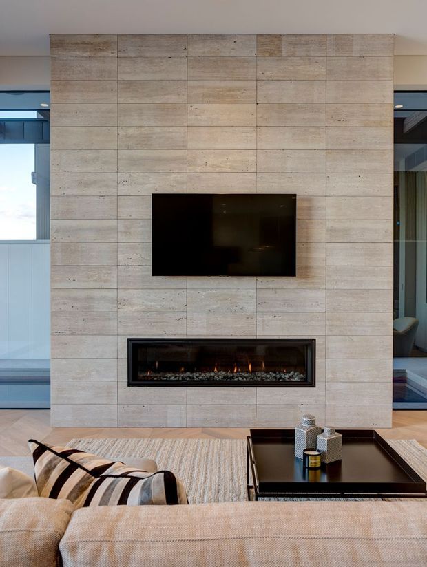 Contemporary living room centered on a full-height pale stone or concrete-look tiled feature wall incorporating a wall-mounted television and long linear fireplace. A large neutral sectional sofa, textured rug, and black coffee table occupy the foreground, with expansive glazing and views into adjacent spaces at either side.