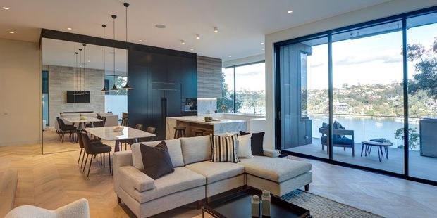 Contemporary open-plan living, dining and kitchen interior with a neutral palette, herringbone timber flooring, dark integrated cabinetry, stone kitchen island, pendant lighting and a large sectional sofa. Full-height glazed sliding doors open to a balcony or terrace with expansive water and hillside views.