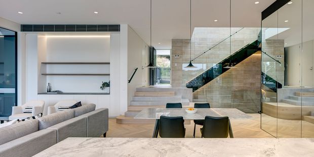 Open-plan contemporary interior combining a living room, dining area and kitchen. A pale stone island or benchtop occupies the foreground, with a light neutral sofa, dining table and black chairs in the middle ground. Timber-toned flooring, white built-in shelving, pendant lights and a dramatic staircase with glass balustrades and mirrored glazing define the space. The staircase wall features large-format beige stone or tile, and greenery is visible through the rear glazing.
