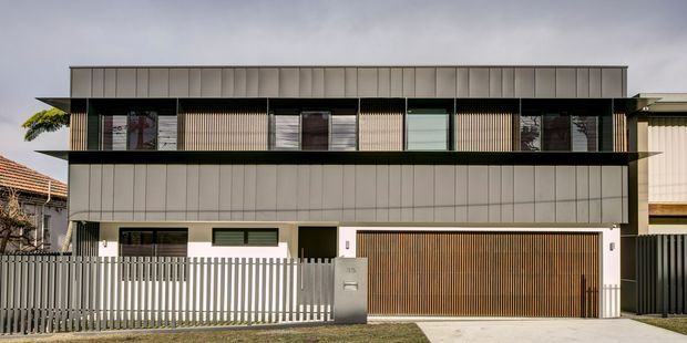 Street-facing view of a contemporary multi-level residence with a strong horizontal composition, pale metal-clad facade, dark recessed upper balcony, vertical screening, and a wide timber garage door. A low metal picket fence and driveway occupy the foreground, with neighbouring residential buildings partially visible at either side.