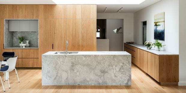 Contemporary open-plan kitchen with a large waterfall-edge stone island, extensive light timber cabinetry, timber flooring, white walls, and integrated dining and living areas. The space features a minimalist, warm-neutral palette with understated lighting and a framed artwork niche.