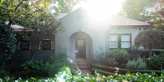 Front exterior view of a single-storey suburban residence partly screened by mature landscaping. The pale rendered facade has a prominent arched entry porch and dark timber door, arched windows to one side, white shuttered windows, and a red tiled pitched roof. Strong backlighting and dense hedges frame the formal garden frontage.