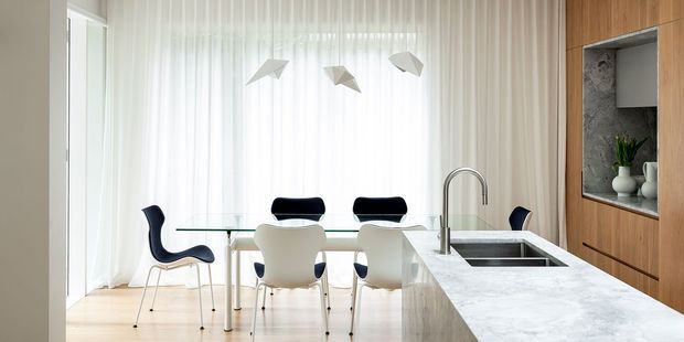 Bright, refined open-plan dining and kitchen area with a glass dining table, sculptural pendant lights, contemporary dining chairs, sheer floor-to-ceiling curtains, and a large white stone kitchen island. Warm timber cabinetry with an integrated display niche and patterned stone backing adds contrast.
