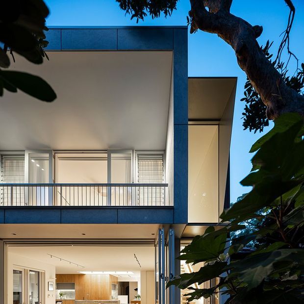 Twilight view of a contemporary multi-storey residence framed by mature tree foliage. The facade features dark blue-grey cladding, crisp projecting volumes, large glazed openings, balconies with slim metal balustrades, and a warmly lit open-plan interior visible at ground level.