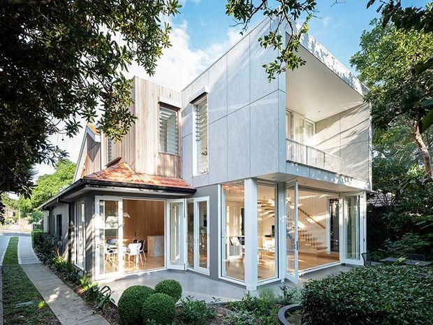 Exterior view of a contemporary multi-storey home combining a restored or retained older wing with a new white-clad extension. The modern addition features extensive floor-to-ceiling glazing, a projecting upper-level balcony, clean geometric forms and an open interior stair visible through the windows. Landscaped garden planting and a paved side walkway frame the house.