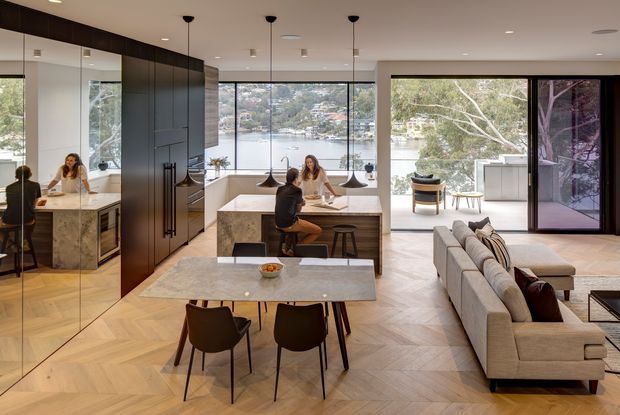 Open-plan contemporary kitchen, dining and living area with expansive floor-to-ceiling glazing overlooking a waterfront setting. The room features dark full-height cabinetry, a large stone kitchen island, marble-look dining table, pendant lighting, herringbone timber flooring, and a neutral sectional sofa. Mirrored cabinetry reflects the kitchen and dining area.