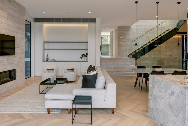Open-plan contemporary living area with a large light-coloured sectional sofa, black metal coffee tables, built-in shelving and a linear fireplace set into pale stone wall cladding. A glass-balustraded staircase rises beside the dining area, while a pale stone kitchen island occupies the foreground. Herringbone timber flooring, recessed lighting and warm concealed shelf lighting create a refined, spacious interior.