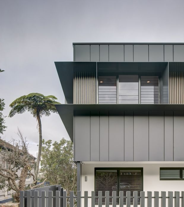 Street-level exterior view of a contemporary multi-storey residence with a sharply cantilevered upper level, dark grey vertical-panel cladding, projecting eaves, and contrasting light rendered walls. The facade includes vertical screening, horizontal window bands, recessed entry glazing, and a grey metal picket fence. Mature landscaping is visible around the property.