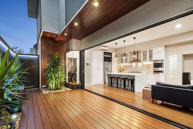 A contemporary open-plan kitchen and living area opening onto a timber-decked outdoor space. The interior features white cabinetry, a light timber floor, a compact island with bar seating, pendant lights, integrated appliances and a dark sofa. The covered deck has timber soffit lining, recessed lighting, vertical timber screening and lush planting.
