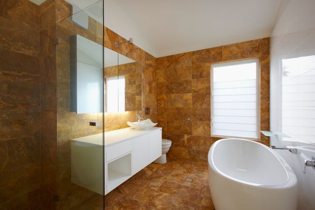 A contemporary bathroom featuring extensive warm brown natural stone or stone-look tile on the walls and floor, a frameless glass shower enclosure, floating white vanity with a vessel basin, toilet, freestanding white bathtub, and bright frosted windows.