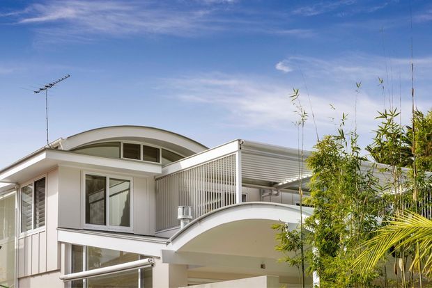 Exterior view of a light-coloured multi-level residence with distinctive curved roof forms, a sweeping arched balcony or canopy, vertical screening, and extensive glazing. Tropical planting partially frames the building, while the bright sky and white facade create an airy contemporary appearance.