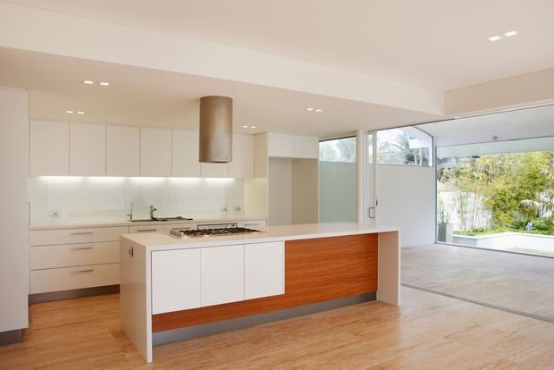 Bright open-plan kitchen with white flat-panel cabinetry, a large island featuring a warm timber front, integrated gas cooktop and cylindrical metal rangehood. The space has light timber flooring, recessed ceiling lighting, pale walls, and extensive glazing opening toward a landscaped outdoor area.