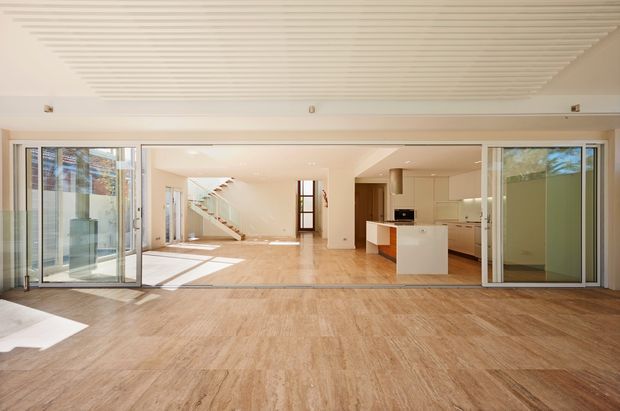 Bright open-plan contemporary interior viewed from a broad glazed opening, with pale timber flooring, a white kitchen island and cabinetry, extensive sliding glass doors, and a staircase leading to an upper level. The space feels airy and connected to the outdoors, with a slatted ceiling adding texture.