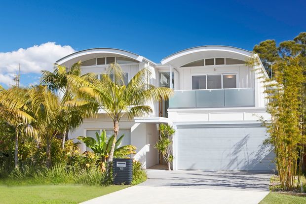 Bright exterior photograph of a contemporary two-storey white residence with two curved roof forms, broad upper-level glazing, a frosted-glass balcony balustrade, and a double garage. Tropical palms and lush garden planting frame the driveway and facade.