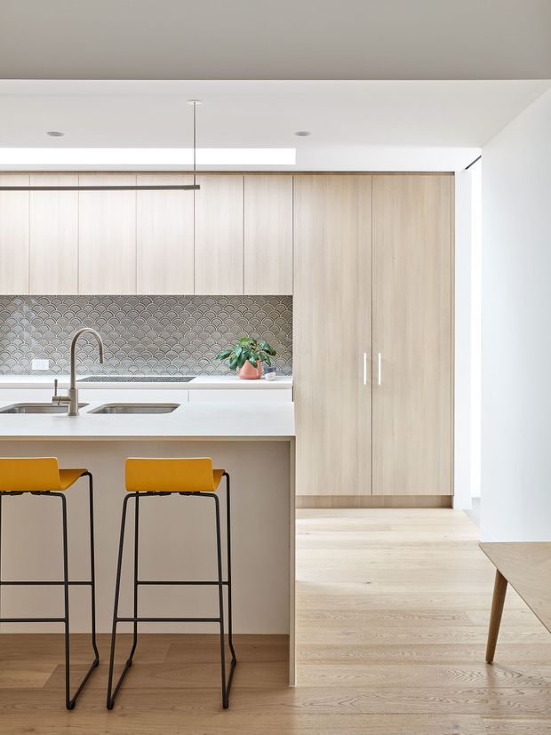 Bright contemporary kitchen with a large pale timber island and matching full-height cabinetry, white benchtops, and a grey scallop-pattern tiled splashback. Two mustard-yellow bar stools sit at the island, with an integrated sink and brushed metal tap. Pale timber flooring extends toward a concealed storage wall and a partially visible adjoining dining or living area. Minimal recessed lighting and a slim linear pendant create a clean, refined interior.