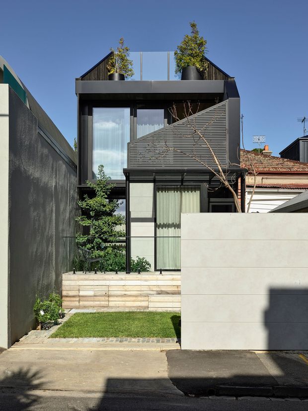 Front exterior view of a compact contemporary multi-level urban residence, showing a dark rendered or metal-clad facade, large glazed openings, slatted screening, rooftop planting, timber terrace steps, and a small lawn enclosed by boundary walls. The house is tightly integrated into a dense suburban streetscape with neighbouring buildings visible at the sides.