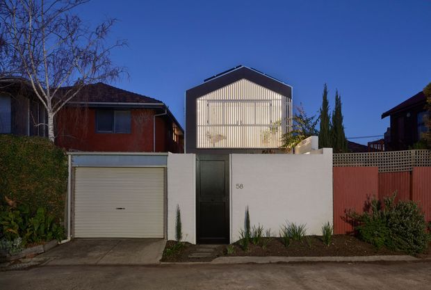 Street-facing view of a compact contemporary residence behind a low white rendered boundary wall. The central upper-level facade has a dark gabled form with vertically screened or slatted glazing, while the frontage includes a dark pedestrian door, an integrated garage with a light roller door, and a red timber boundary fence. Established planting and neighbouring residential buildings frame the property.