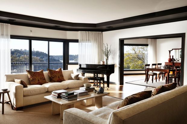 A bright, spacious living room with floor-to-ceiling glazing overlooking a leafy landscape and water, furnished with cream sofas, a substantial stone or light-toned coffee table, patterned cushions, and a black grand piano. A wide opening connects the living room to a dining area with timber furniture. The room features dark recessed ceiling detailing, large black-framed windows and doors, sheer curtains, and warm natural light.