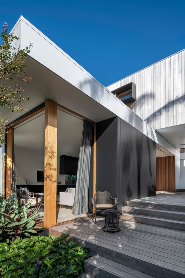 Exterior view of a contemporary multi-level home with crisp white geometric roof forms, vertical light cladding, dark metal wall panels, and extensive timber decking and steps. Large glazed openings reveal a living room, while a timber post, outdoor chairs, lush planting, and strong sunlight create a connection between the interior and landscaped deck.