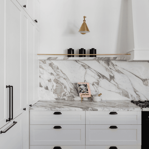 A refined white kitchen with full-height shaker-style cabinetry, black hardware, a dramatic white-and-grey veined stone benchtop and matching splashback, brass wall light, and a slim open display shelf with dark canisters. A cooktop is partially visible at right.