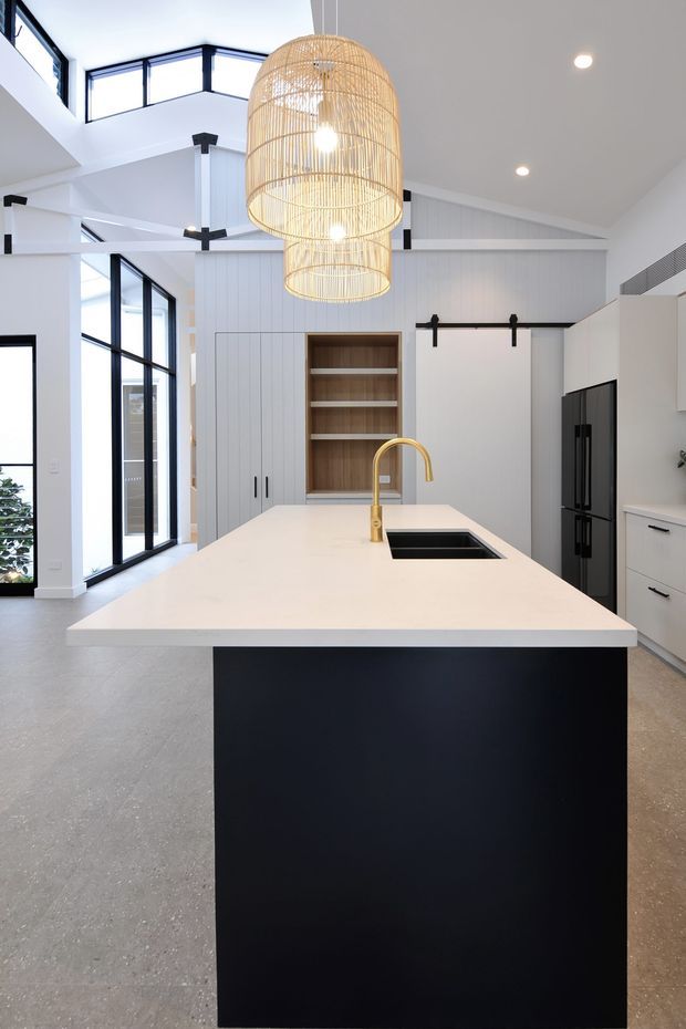 Bright contemporary kitchen with a large white island, dark cabinetry, integrated black sink, and brass tap. The space features tall black-framed glazing, white vertical wall panelling, exposed pitched ceiling lines, a barn-style sliding door, and a large woven pendant light over the island.