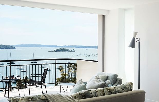 Bright contemporary living room with a large sectional sofa, patterned cushions, a slim black floor lamp, and a small outdoor-style table and chairs near full-height glazing. The room opens onto a balcony with a black metal balustrade and overlooks a calm bay with boats and wooded shoreline.