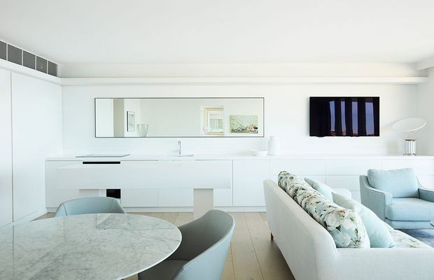 Bright open-plan contemporary living and kitchen space with predominantly white built-in cabinetry, a pale stone kitchen island, light timber flooring, a round dining table with upholstered chairs, and a light sofa and armchair. A large horizontal mirror and wall-mounted artwork add visual interest, while concealed linear air conditioning runs along the ceiling.