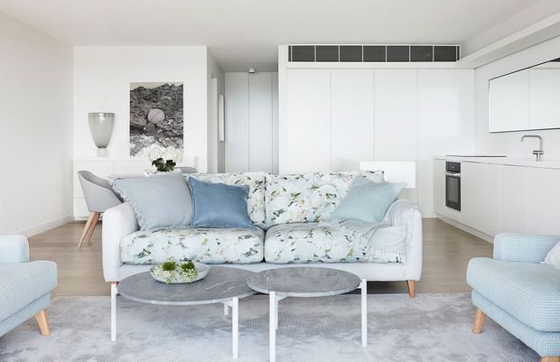 Bright open-plan living room with a white floral-patterned sofa, pale blue accent cushions, two round nesting coffee tables, and matching pale blue armchairs. A minimalist white kitchen with integrated cabinetry, built-in oven, and island or peninsula is visible behind the seating area. The room has light timber flooring, white walls and cabinetry, soft grey carpeting, and understated contemporary decor.