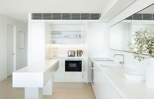 Bright, minimalist white kitchen with a compact island, integrated oven, open shelving, white cabinetry, a long stone-look benchtop, undermount sink, and a large wall mirror reflecting the space. Pale timber flooring and concealed linear ventilation add subtle contrast.
