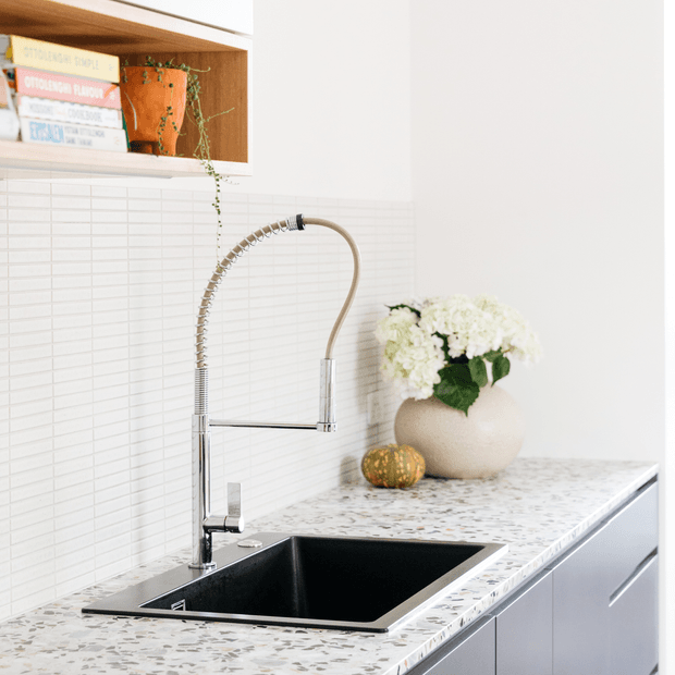 Bright contemporary kitchen detail featuring a black undermount sink with a high-arched spring-style chrome tap, pale terrazzo-look benchtop, white narrow rectangular tiled splashback, and light grey cabinetry. Open timber shelving displays books and a trailing potted plant, while a vase of white flowers adds a soft decorative accent.