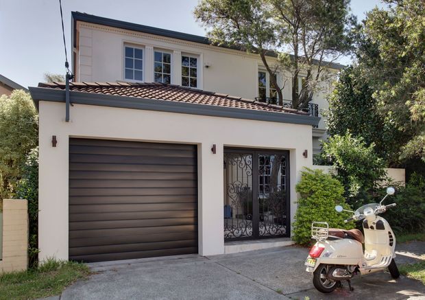 Street-level exterior view of a two-storey residential building with a cream rendered facade, dark tiled low-pitched roof, enclosed single-car garage, and ornate wrought-iron entry gates. Mature landscaping partially frames the building, and a scooter is parked in the driveway.