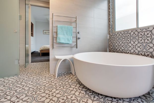 A bright contemporary bathroom featuring a freestanding white oval bathtub, wall-mounted chrome towel warmer, pale tiled walls, and patterned encaustic-style floor and partial wall tiles. A bedroom is visible through the open doorway.