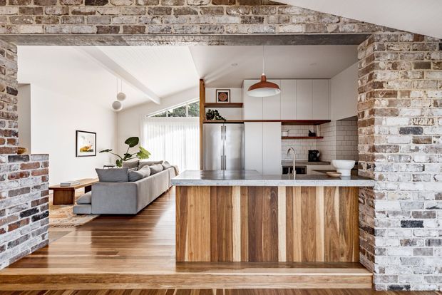 Open-plan kitchen and living area viewed through a wide exposed brick opening. The kitchen features a timber-clad island with a light benchtop, white flat-panel cabinetry, stainless-steel appliances, and a white tiled splashback. A grey sectional sofa and timber flooring extend into the adjacent living space. White ceilings, exposed brick walls, pendant lighting, and a split-level timber step define the interior.