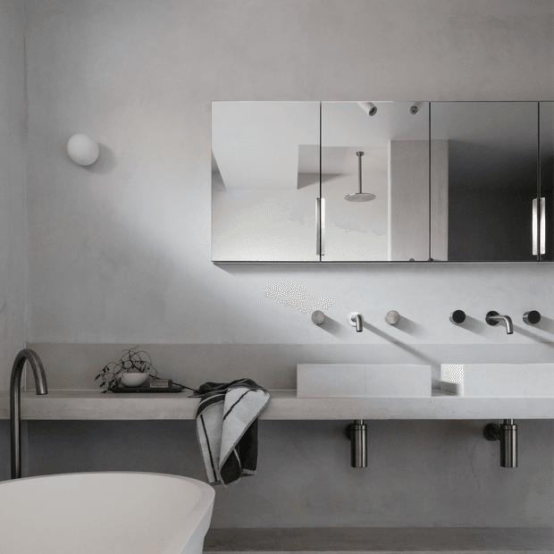 A minimalist contemporary bathroom with a long pale stone double vanity, two integrated rectangular basins, wall-mounted chrome taps, exposed cylindrical plumbing, and a large mirrored cabinet. A freestanding white bathtub and black-framed faucet occupy the foreground. The room has smooth grey plaster walls, restrained monochrome styling, and a wall sconce.