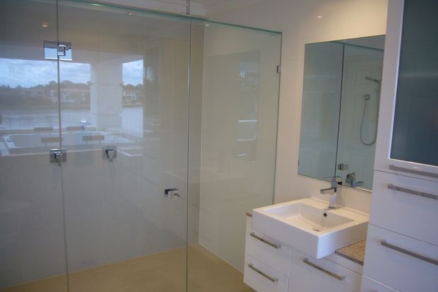 A contemporary bathroom featuring a large frameless glass shower enclosure with chrome hardware, a wall-mounted white vanity with integrated basin and drawers, a large mirror, and pale neutral wall and floor finishes. A window provides a view toward water and greenery outside.