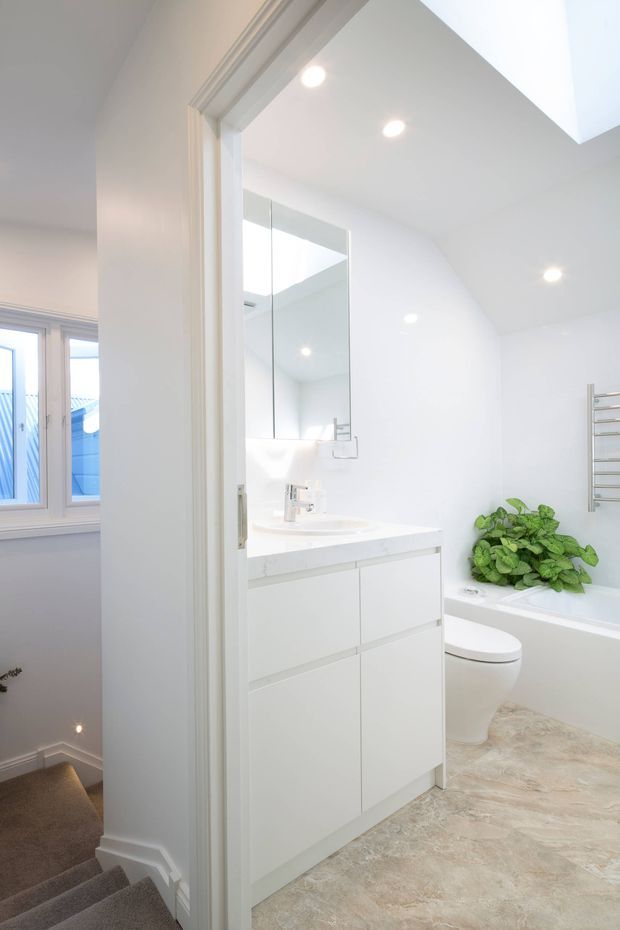 Bright, compact contemporary bathroom viewed through an open doorway, with a white wall-mounted vanity, integrated basin, large mirrored cabinet, bathtub, toilet, heated towel rail, recessed ceiling lights, and pale stone-look flooring. A green plant adds contrast. A carpeted stairway and window are visible outside the bathroom.
