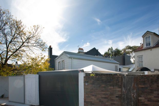 Exterior view of a compact residential property behind a boundary wall and driveway gate. The main building is a light-painted, horizontal-clad single-storey structure with a pitched roof, chimney, and small roof-level additions. Adjacent older masonry homes and mature trees are visible, with a bright blue sky and strong sunlight.