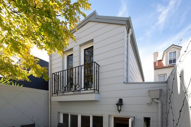 Exterior view of a compact two-storey white-clad building in a courtyard setting, featuring horizontal weatherboard-style siding, tall windows, a small projecting balcony with ornate black metal balustrade, simple boxed roof forms, and traditional exterior wall lighting. A neighbouring period-style building is visible beyond the courtyard wall.