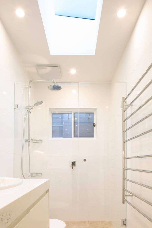Bright, compact contemporary bathroom with a frameless glass shower enclosure, wall-mounted shower fittings, white tiled walls, a skylight, recessed ceiling lights, a small window, a white vanity with pale stone benchtop, and a stainless-steel heated towel rail. A partial bathtub and toilet are visible at the lower edge.