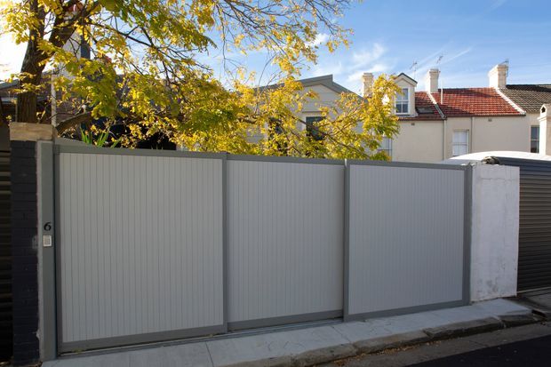 Street-facing view of a contemporary residential property boundary, dominated by a tall grey metal pedestrian and vehicle gate with vertical ribbed panels and dark posts. A house with traditional pitched roofs, chimneys and red terracotta roof tiles is visible behind the boundary, along with mature trees and neighbouring urban residential buildings.