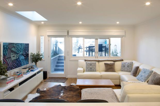 Bright, contemporary living room with a large cream sectional sofa, timber coffee table, textured neutral cushions, and a low white media/storage unit. A colourful abstract artwork and stacked books add visual interest. The room has recessed ceiling lights, white walls and trim, a skylight, and wide windows and a glazed door opening toward an exterior stair and patio area.