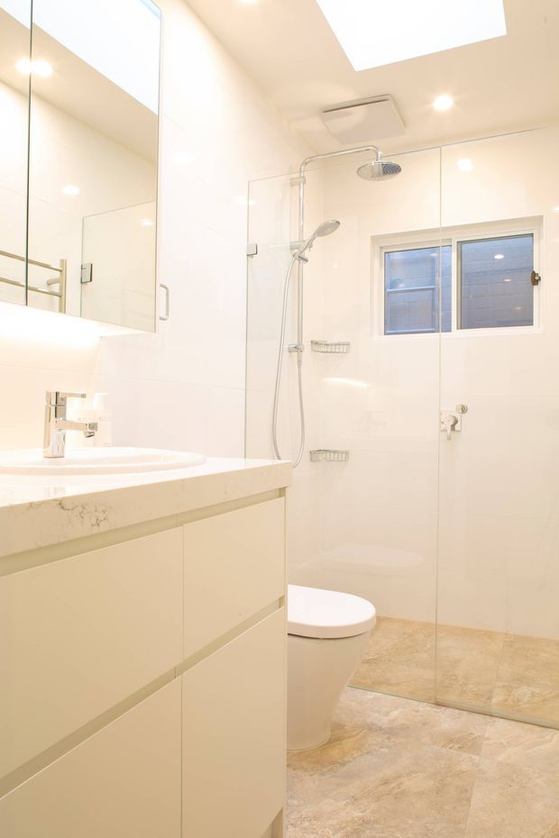 Bright, compact contemporary bathroom with a wall-mounted vanity, white integrated basin, chrome tapware, large mirrored cabinet, glass-enclosed walk-in shower, toilet, small window, recessed ceiling lights, and a skylight. The room uses predominantly white finishes with warm beige stone-look floor tiles and a pale stone benchtop.