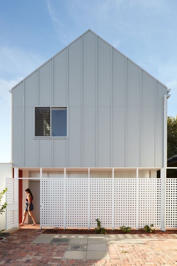 Front exterior view of a contemporary two-storey gabled house. The upper facade is clad in pale grey vertical panels with expressed seams and a small horizontal window, while the lower level is screened by white breeze-block masonry and a covered entry walkway. A patterned brick and concrete-paver forecourt occupies the foreground.