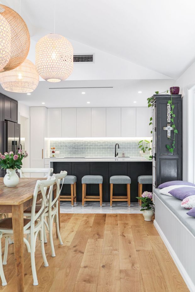 Bright open-plan kitchen and dining area with white cabinetry, a dark island base, pale brick-tile splashback, timber flooring, and grey upholstered bar stools. A timber dining table with white chairs occupies the foreground, while woven pendant lights, indoor plants, and a built-in window seat add warmth and texture. The room has a sloped white ceiling and contemporary coastal-inspired styling.