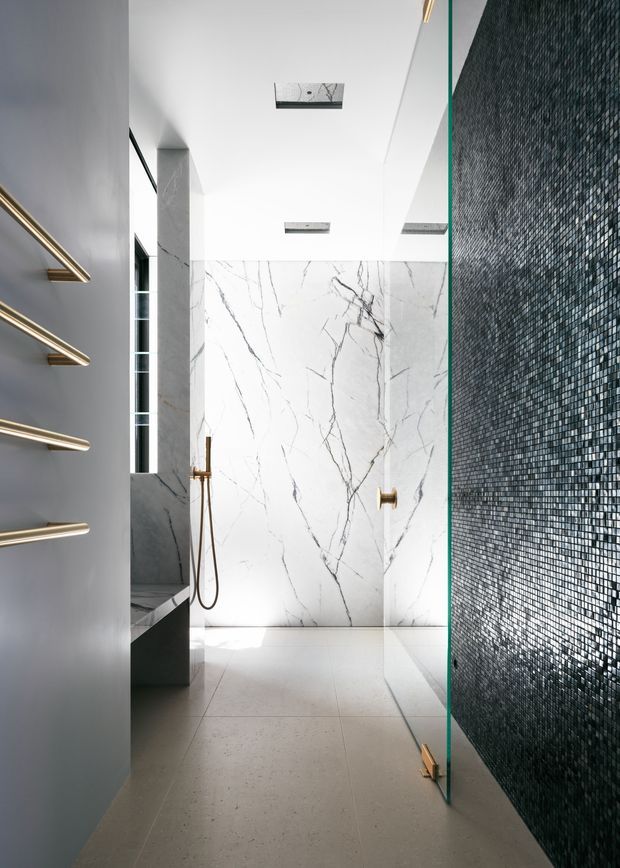 A refined contemporary bathroom with a walk-in shower, large white marble-look wall slabs with dramatic grey veining, pale stone floor tiles, a dark textured mosaic-tile feature wall, frameless glass shower screen, and brushed brass shower and towel hardware. Recessed ceiling lights and a narrow vertical window provide bright natural illumination.