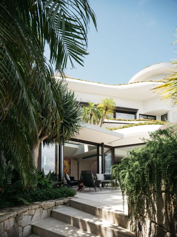 A bright contemporary multi-level residence surrounded by lush tropical landscaping. The white sculptural facade features sweeping curved rooflines, broad glass openings with dark frames, layered terraces, and an outdoor seating area accessed by broad stone steps. Dense palms, hedges, and planted retaining walls frame the courtyard-like exterior.