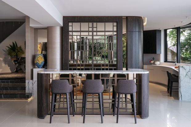 Contemporary open-plan interior featuring a dark timber and metal-framed bar or island with three upholstered stools, a large decorative grid-framed mirror, curved timber-clad columns, polished light flooring, and an adjacent kitchen with pale cabinetry and a marble-patterned island. A stairway and decorative planting are visible to the left.