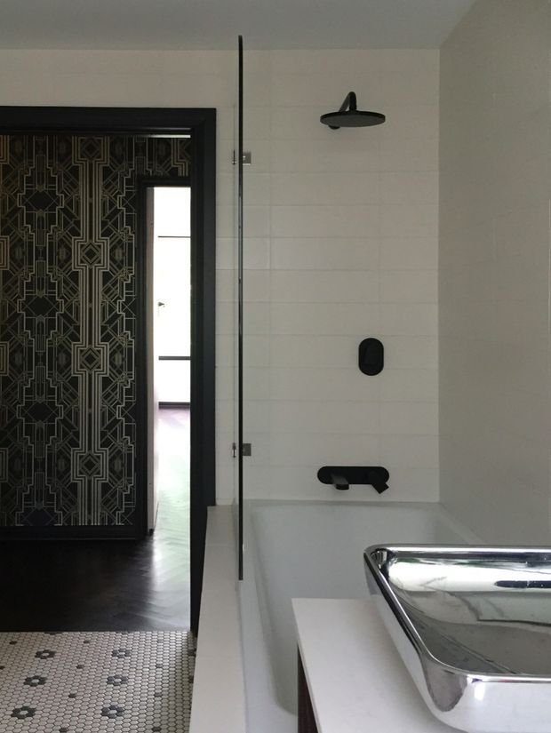 A contemporary bathroom with a white tiled shower wall, frameless glass shower screen, matte black showerhead and controls, and a freestanding or inset white bathtub. A polished chrome basin is visible in the foreground. The room includes a striking black geometric patterned door and dark timber flooring beyond the bathroom threshold, creating a strong monochrome contrast.