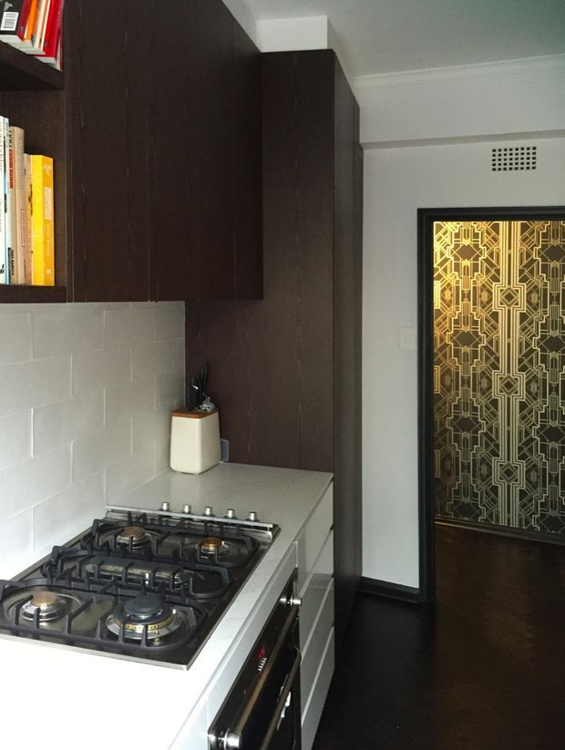 A compact kitchen viewed toward a doorway. The space features a white countertop with a built-in four-burner gas cooktop and oven, pale rectangular tiled splashback, dark timber overhead and tall cabinetry, and dark flooring. A black-framed doorway leads to a room with an ornate geometric patterned glass panel. Bookshelves are visible at the upper left, and the kitchen has a restrained, high-contrast palette with a decorative art-deco-like doorway detail.